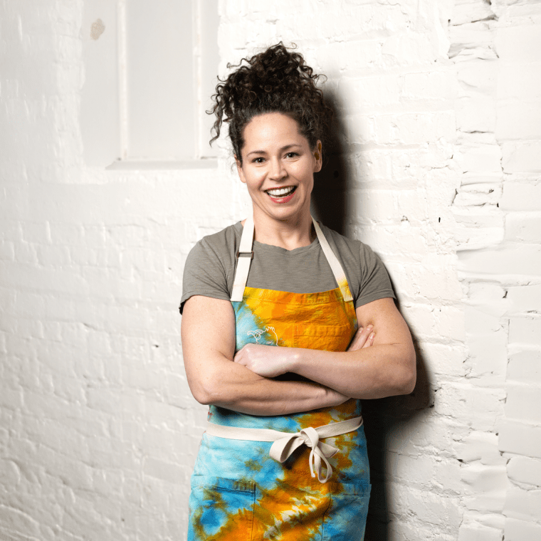 Stephanie Izard in a blue and gold apron leaning agains a white brick wall