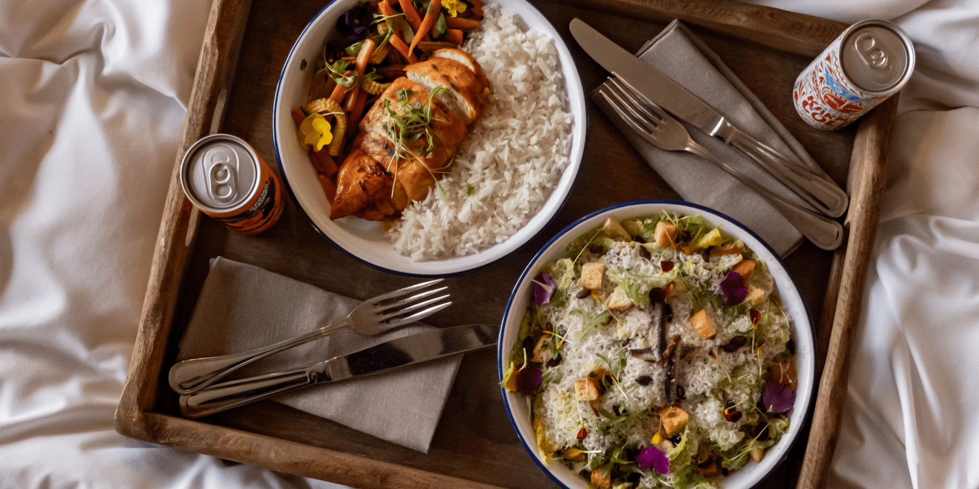 Two meals sit on a wooden tray with linen napkins on a bed at Treehouse hotels