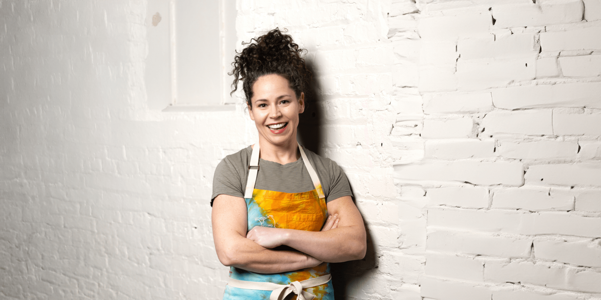 Stephanie Izard in a blue and gold apron leaning agains a white brick wall