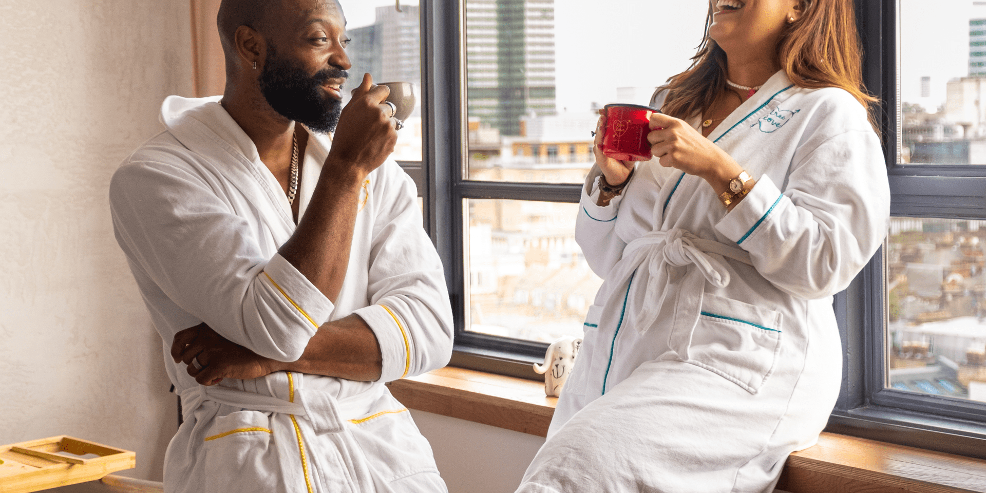 A couple wearing bathrobes laugh while leaning up against a hotel room window and drink coffee.