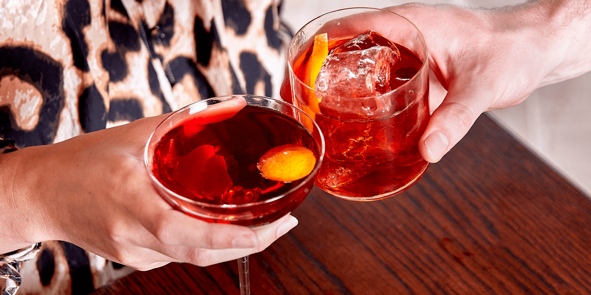 Two hands clinking glasses of cocktails at a bar.