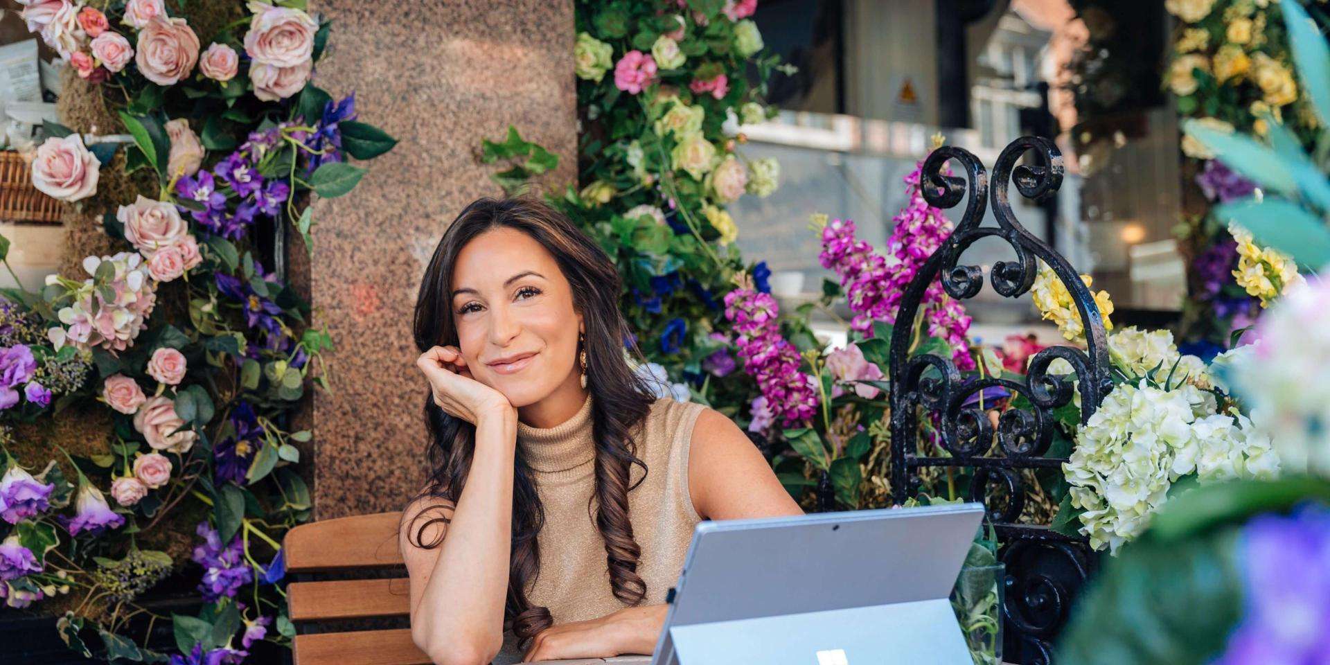 Jacqueline Hurst sitting at a wood table on her laptop surrounded by flowers