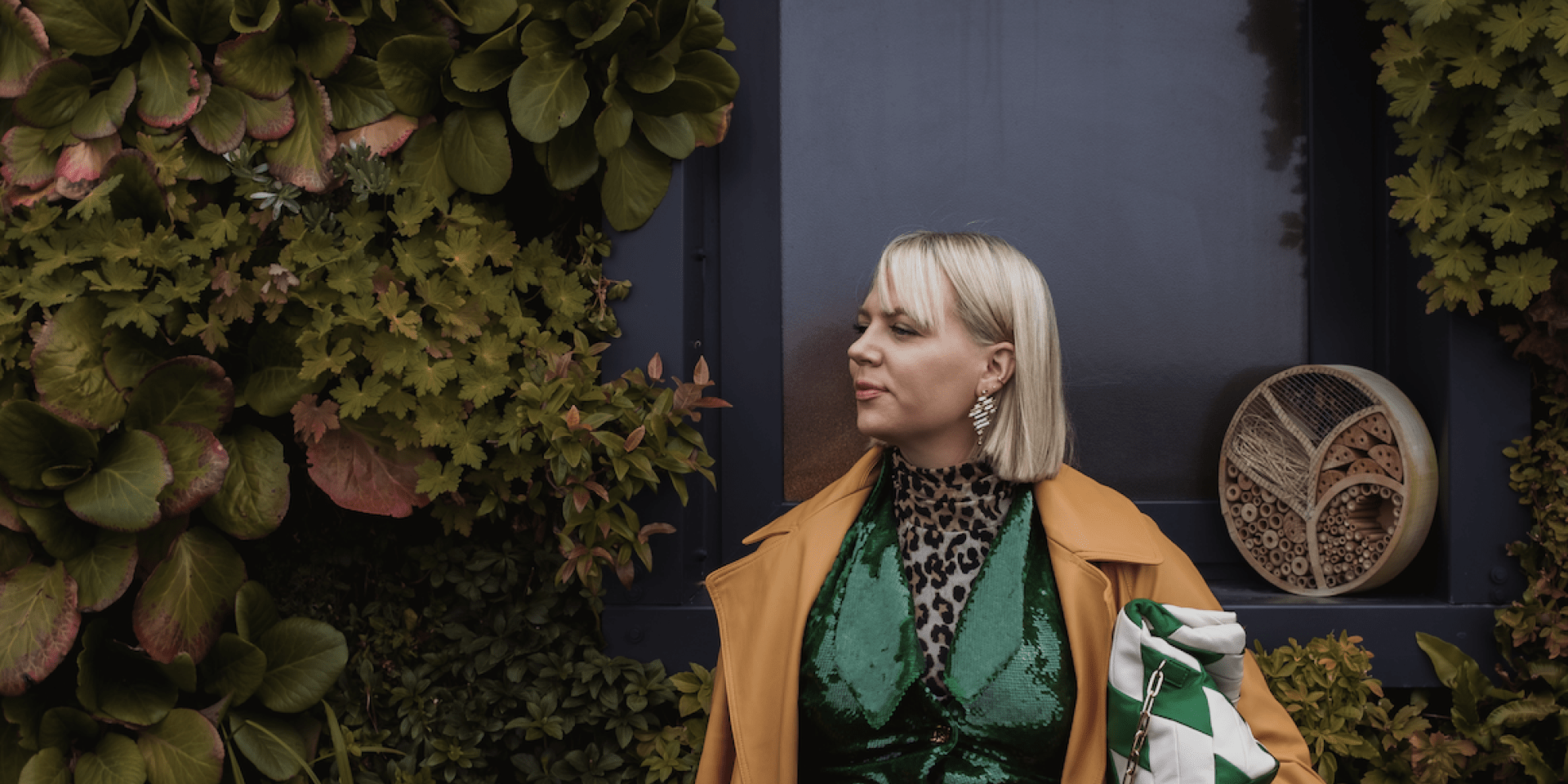 Cassandra Dittmer, a woman with blond short hair and fashionable clothing, stands in front of a lush green space.
